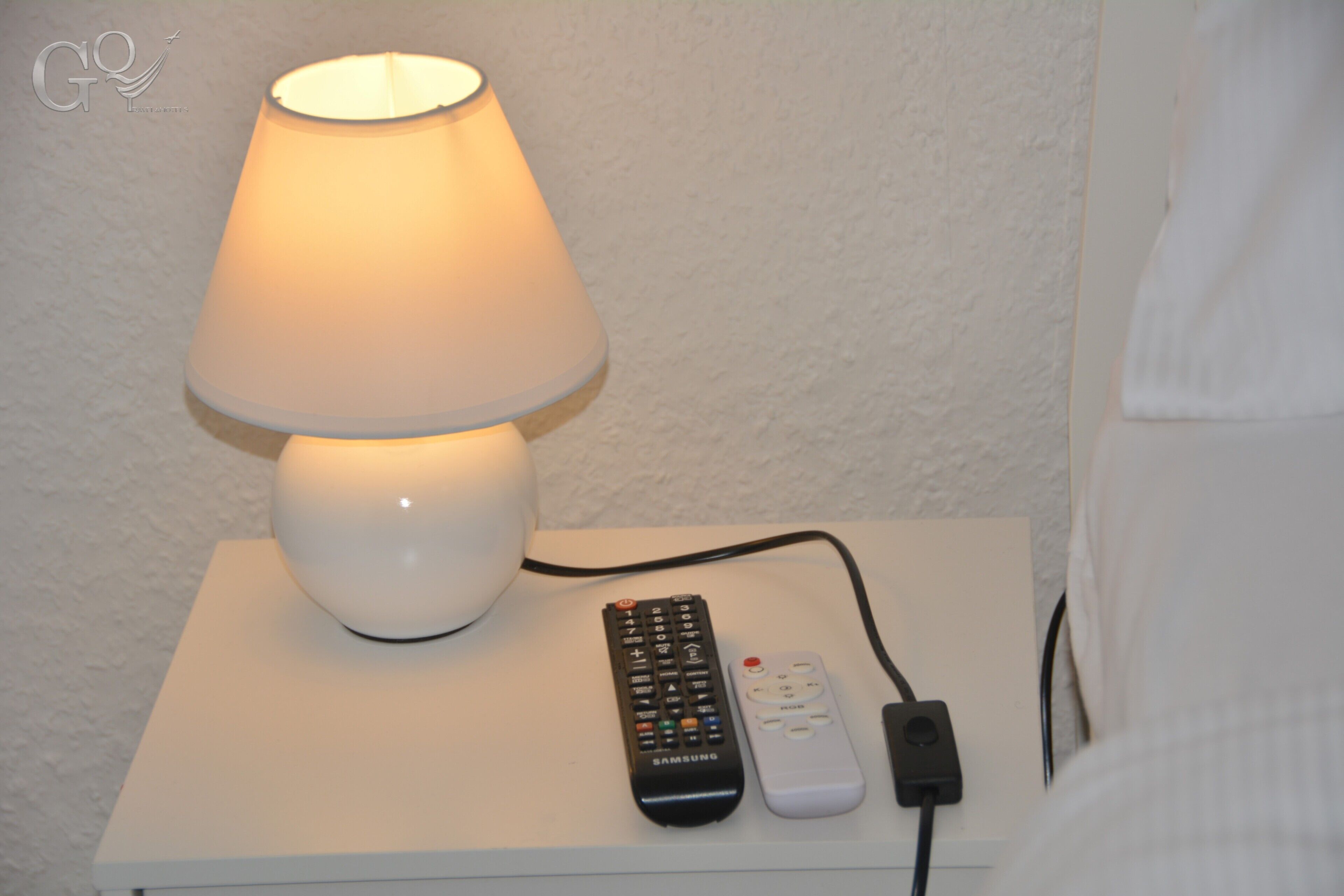 A white bedside table with a lamp, a remote control, and a power adapter.