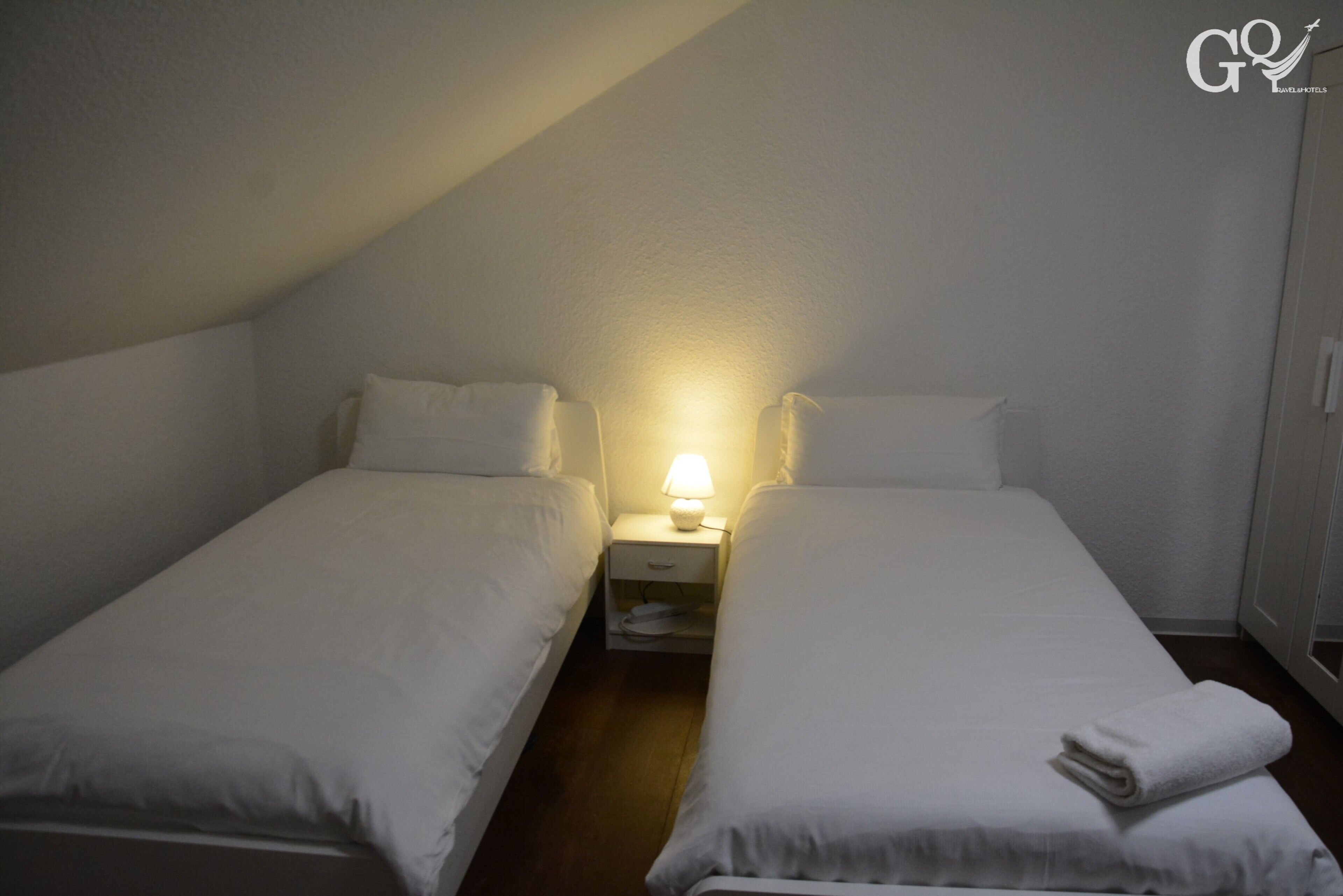 Habitació petita i poc il·luminada amb dos llits individuals, una tauleta de nit amb una làmpada i una paret blanca.