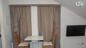 Quarto com uma mesa branca e duas cadeiras de vime, uma TV fixada na parede e cortinas com padrão circular.