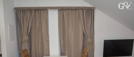 A room with a white table and two wicker chairs, a TV mounted on the wall, and curtains with a circular design.