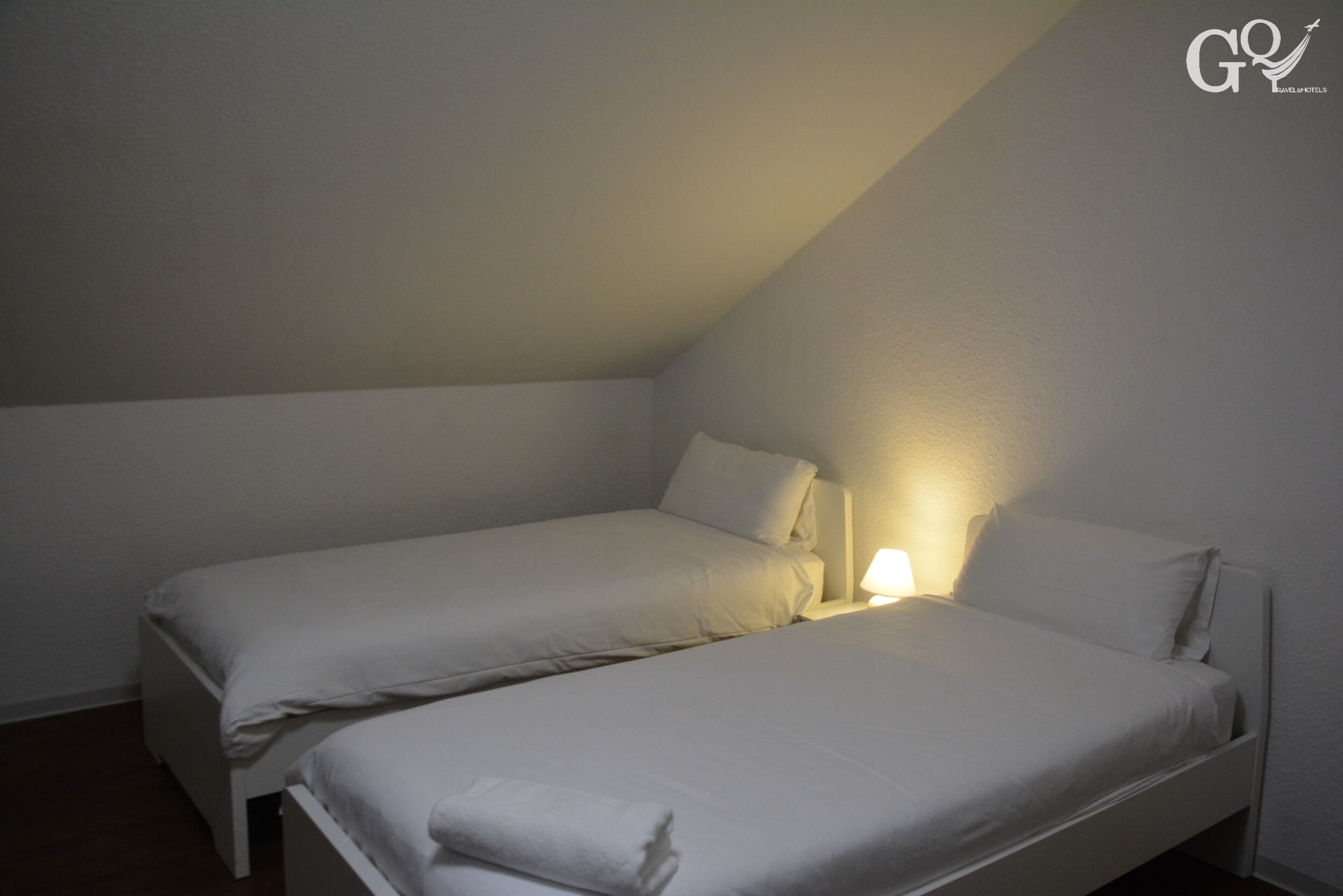 A small, dimly lit room with two single beds, a nightstand with a lamp, and a sloped ceiling.
