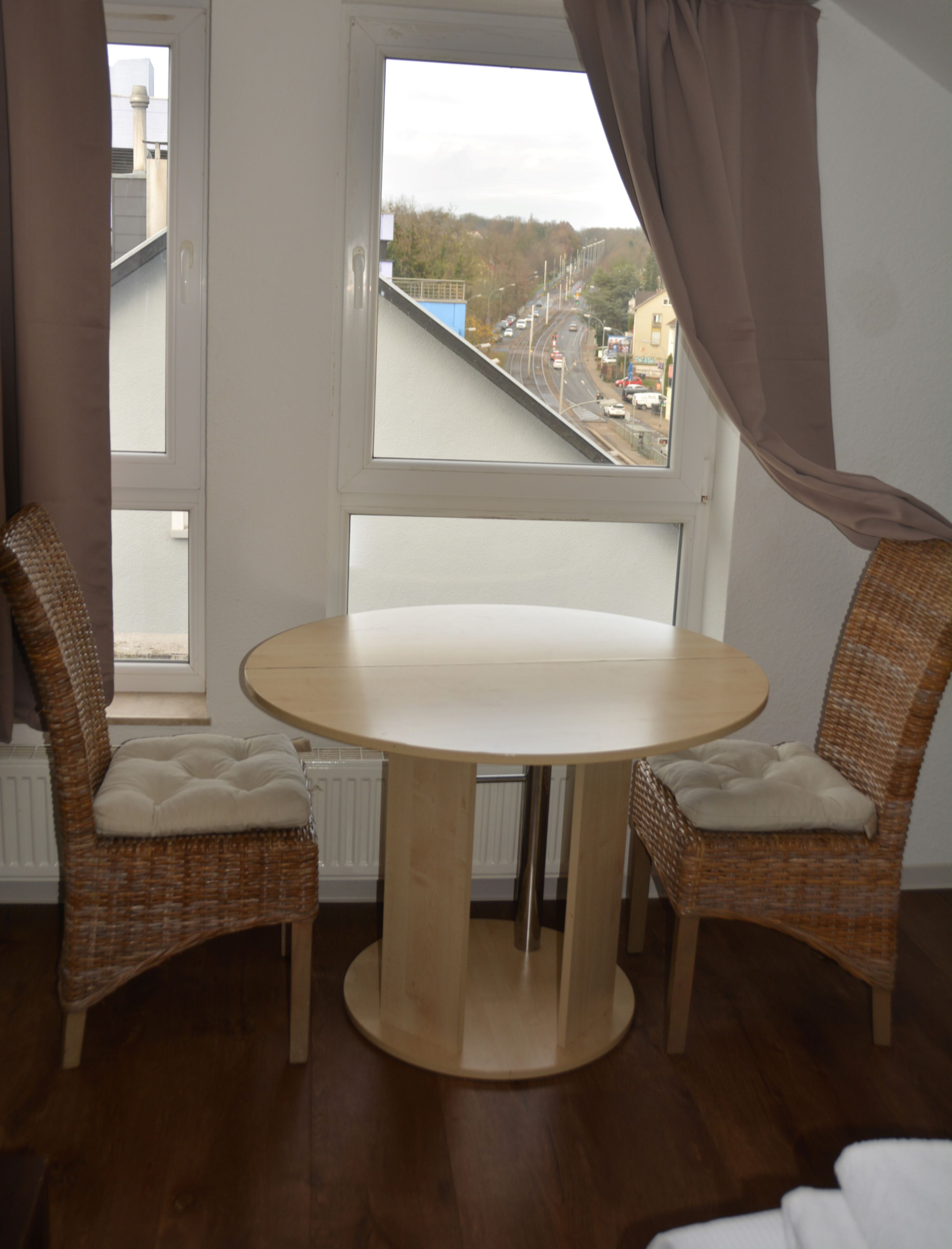 Une pièce avec une table ronde et deux chaises en osier dotées de coussins, une fenêtre avec des rideaux et une vue sur une route et des bâtiments à l’extérieur.