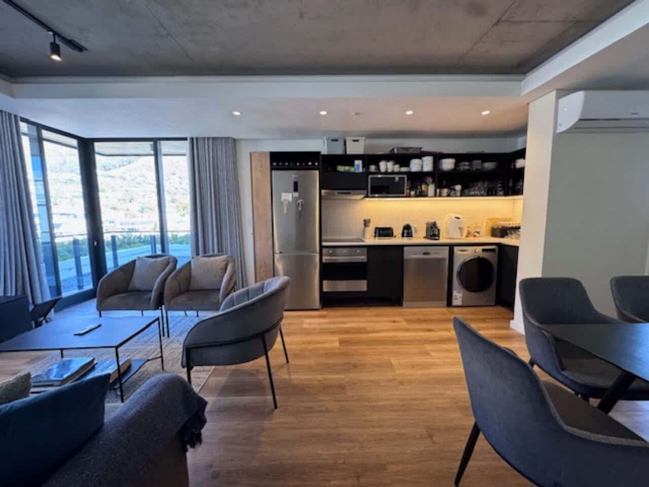 Ein modernes Wohnzimmer mit einer Couch und Sesseln, einem Esstisch und einer Küchenzeile mit Geräten.