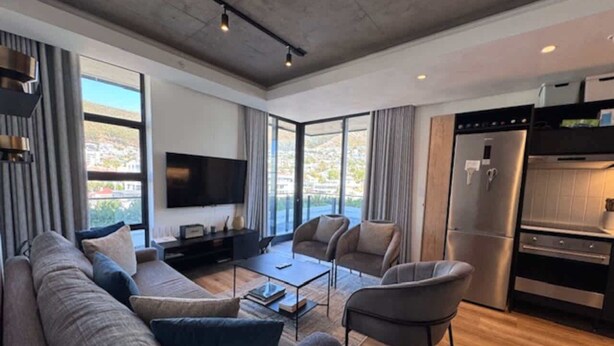 Ein modernes Wohnzimmer mit einer Couch, Sesseln, einem Couchtisch und einem Flachbildfernseher. Das Zimmer verfügt über große Fenster und einen Küchenbereich mit Kühlschrank und Backofen.