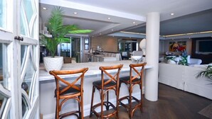 A spacious living area with a kitchen, a dining area with three wicker chairs, and a large potted plant.