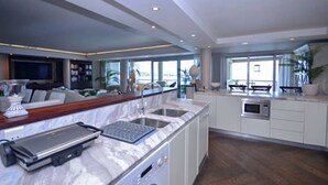 A modern kitchen with a marble countertop, a built-in oven, and a sink. There is a dining area with a table and chairs, and a large window providing a view of the outdoors.
