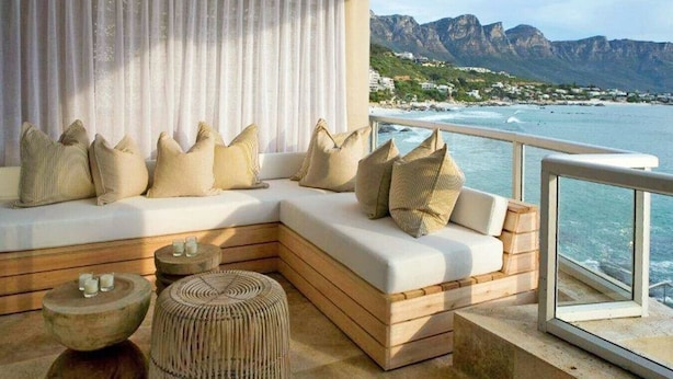A balcony with a white cushioned sofa, beige cushions, a woven stool, and a view of the ocean and mountains.