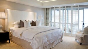A spacious bedroom with a large bed, a nightstand, a chair, and a view of the outside through large windows.