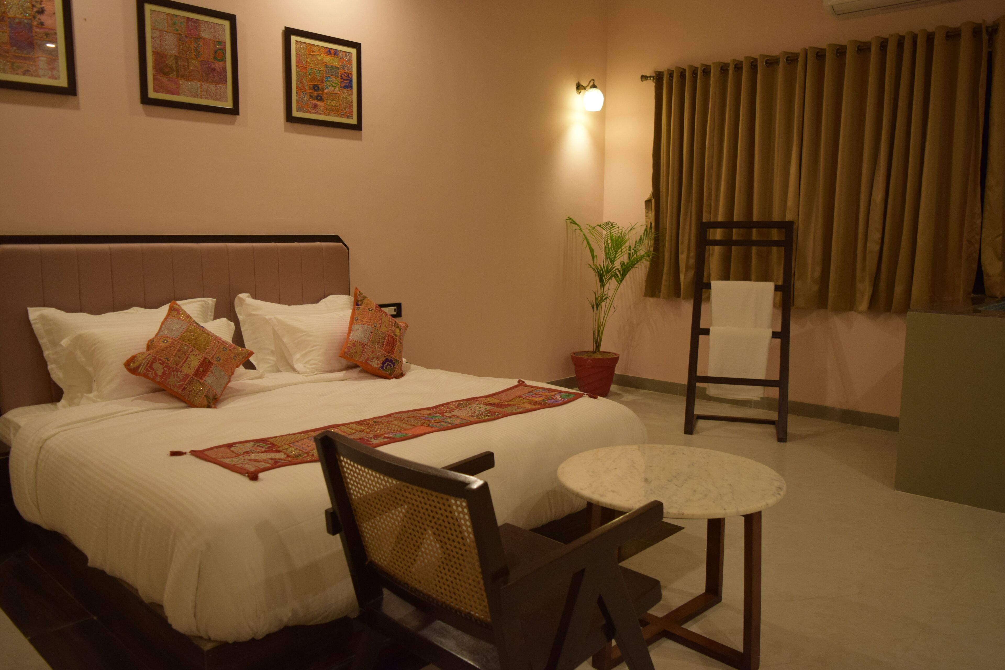 A neatly made bed with white linens and decorative pillows, a small round table, a chair, a potted plant, and a wall-mounted light fixture.
