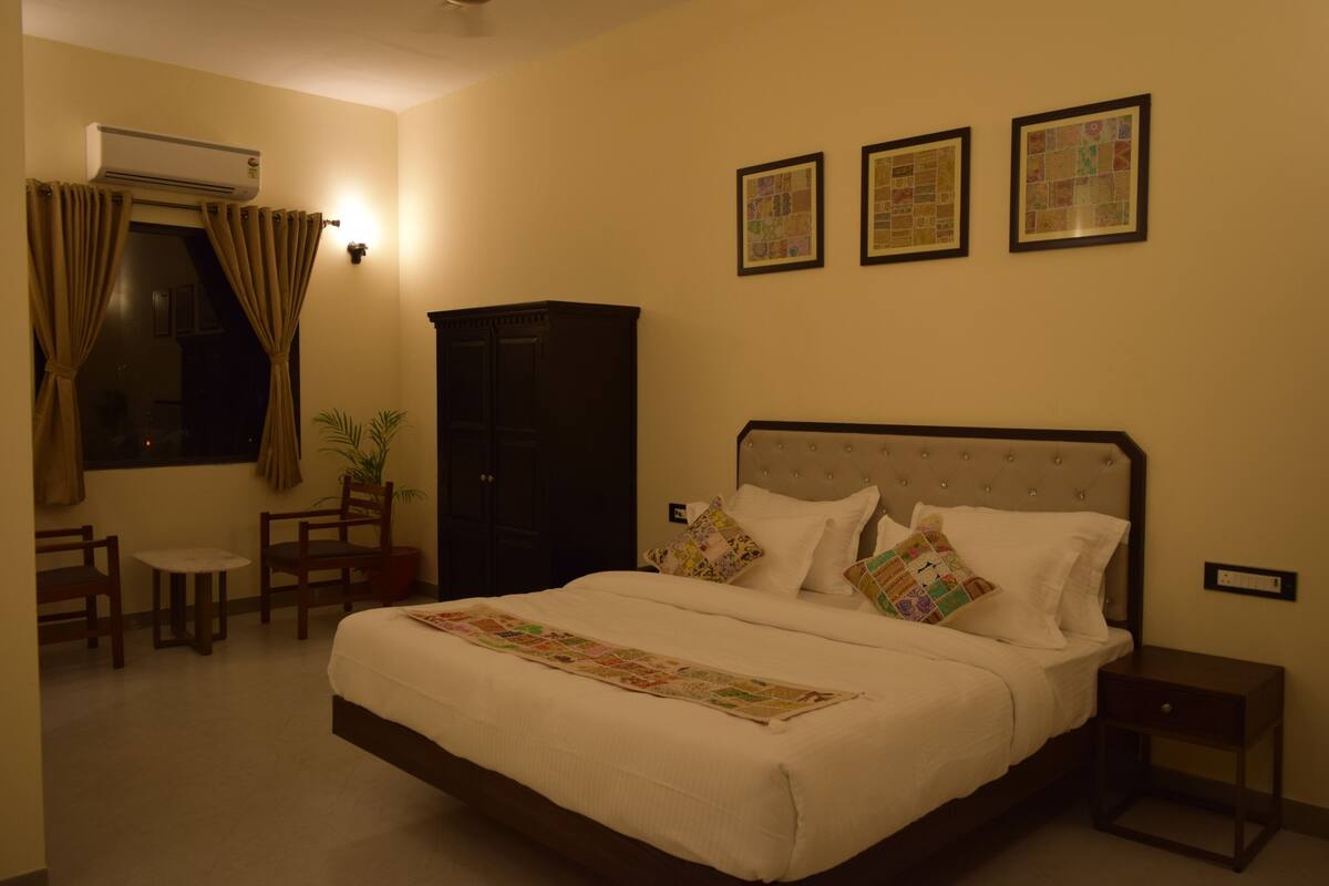 A hotel room with a large bed, a small table, two chairs, a wardrobe, and three framed pictures on the wall.