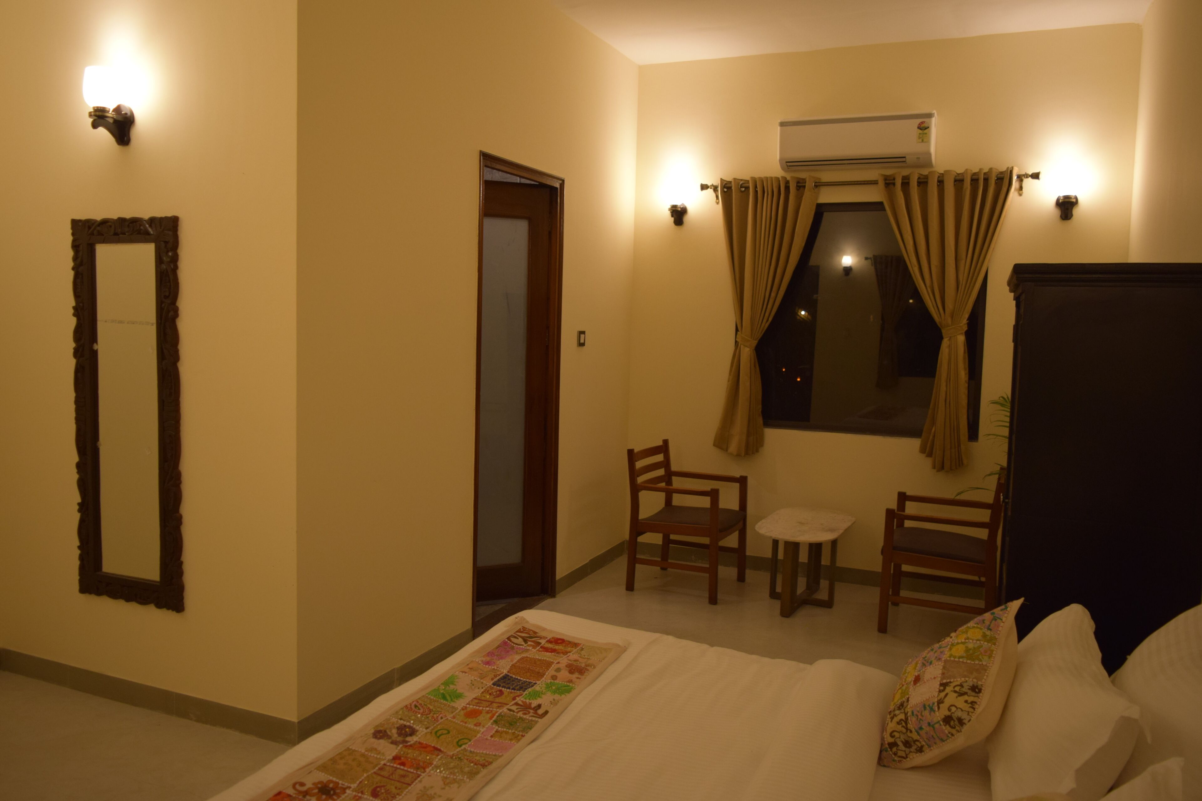 A hotel room with a bed, two chairs, a small table, a mirror, and an air conditioning unit.