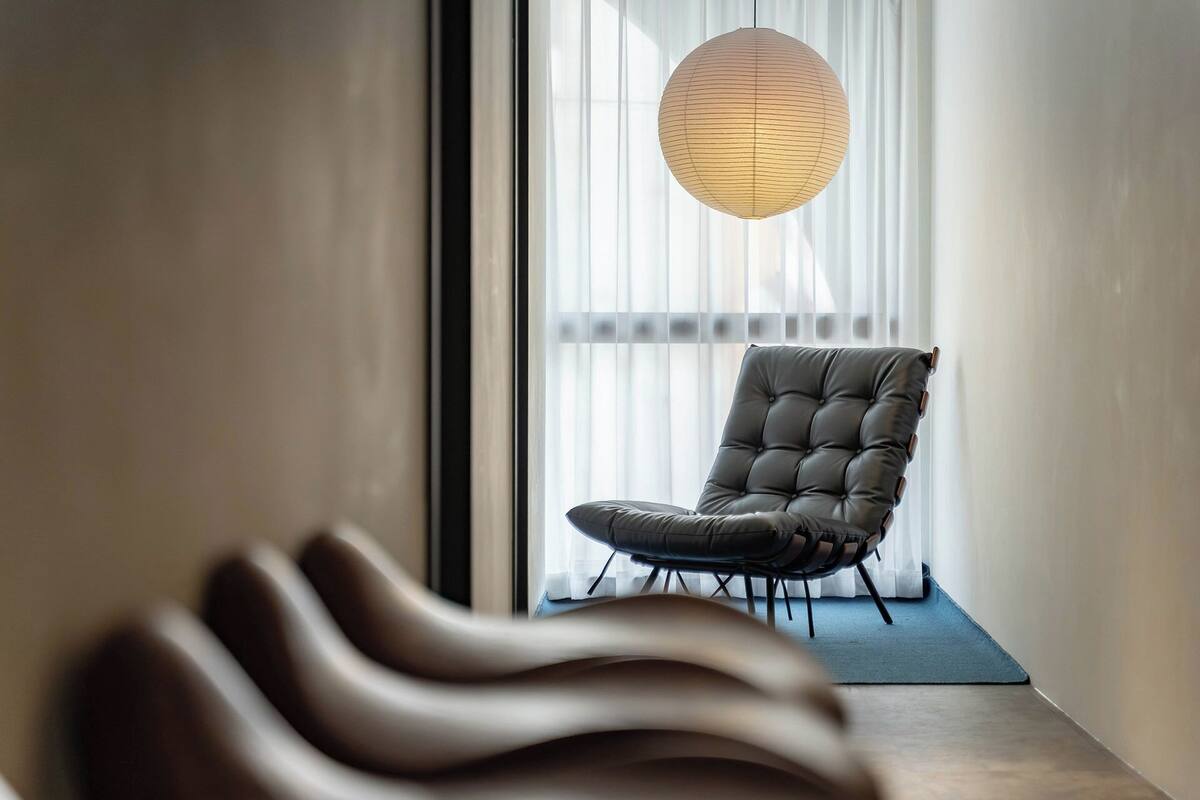 A modern interior with a tufted leather chair, a hanging paper lantern, and a view through sheer curtains.