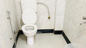 A small bathroom with a toilet, a shower, and a plastic container on a stool. - Hotel Churu Palace (Jaipur)