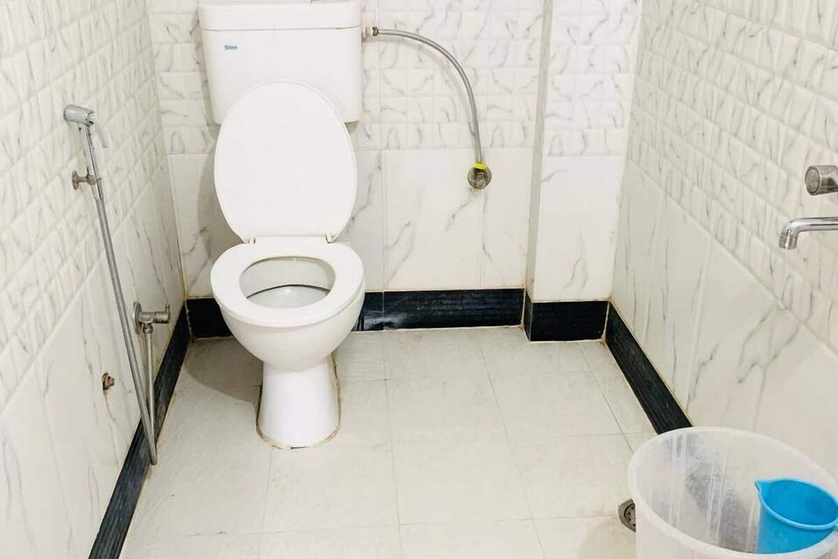 Une petite salle de bain avec des toilettes, une douche et un récipient en plastique posé sur un tabouret.