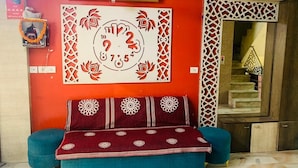 A room with a sofa set against a red wall, a decorative wall piece, and a staircase in the background. - Hotel Churu Palace (Jaipur)