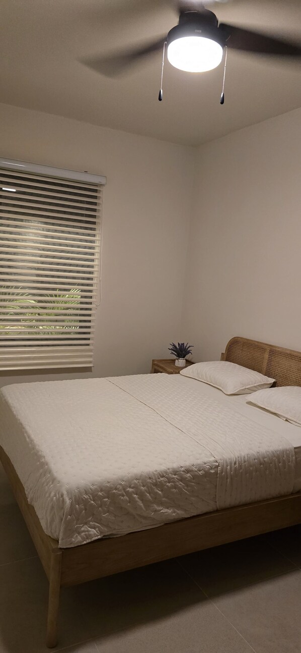 A bedroom with a bed, a ceiling fan, a nightstand with a plant, and a window with blinds.