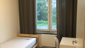 A small, single-bed room with a wooden frame, a white desk, and a window with curtains. - Hallera Wroclaw (Wrocław)