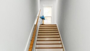 Een trap met houten treden en witte muren, die leidt naar een kamer met een raam en een blauwe vaas.