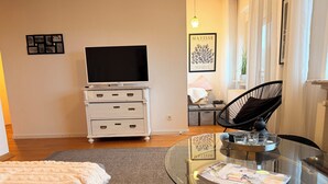 Ein modernes Wohnzimmer mit einem Glastisch, einer Couch, einer weißen Kommode mit Schubladen, einem Flachbildfernseher und einem gerahmten Kunstwerk an der Wand.