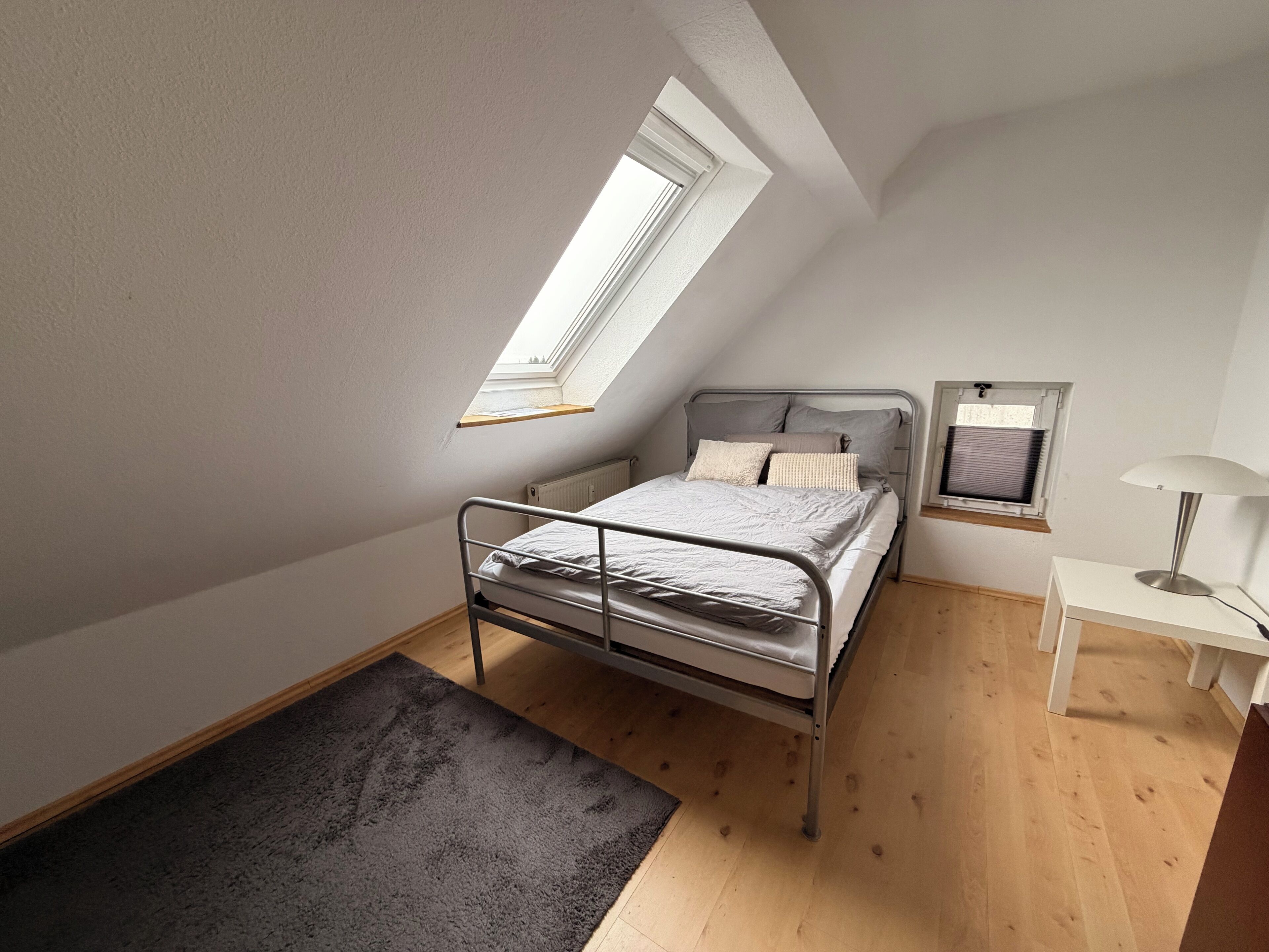 Une petite chambre à coucher, bien rangée, avec un plafond en pente, un lit simple, une table de chevet, une lampe et une fenêtre.
