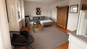 Ein modernes Wohnzimmer mit grauem Sofa, einem Glastisch, einem Korbstuhl und einem Holzschrank.