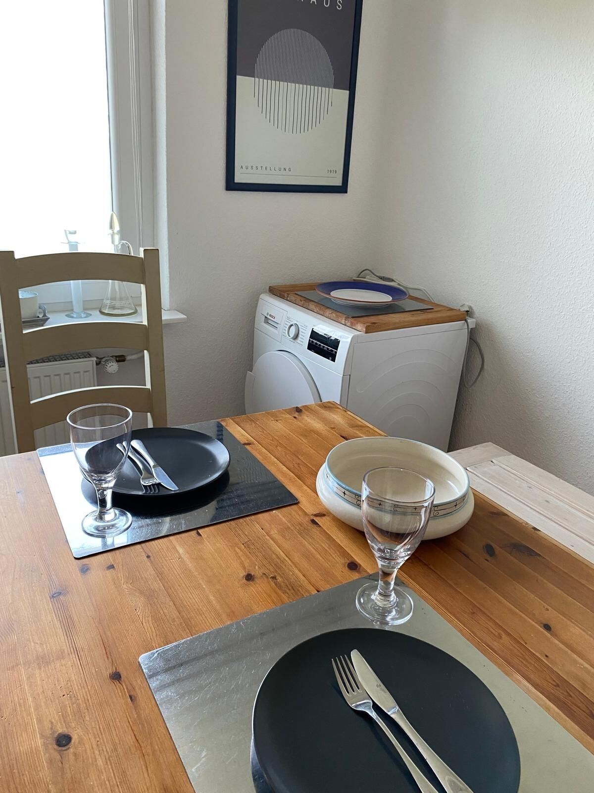 Une table à manger dressée avec des assiettes, des couverts et des verres, un lave-linge et une chaise.