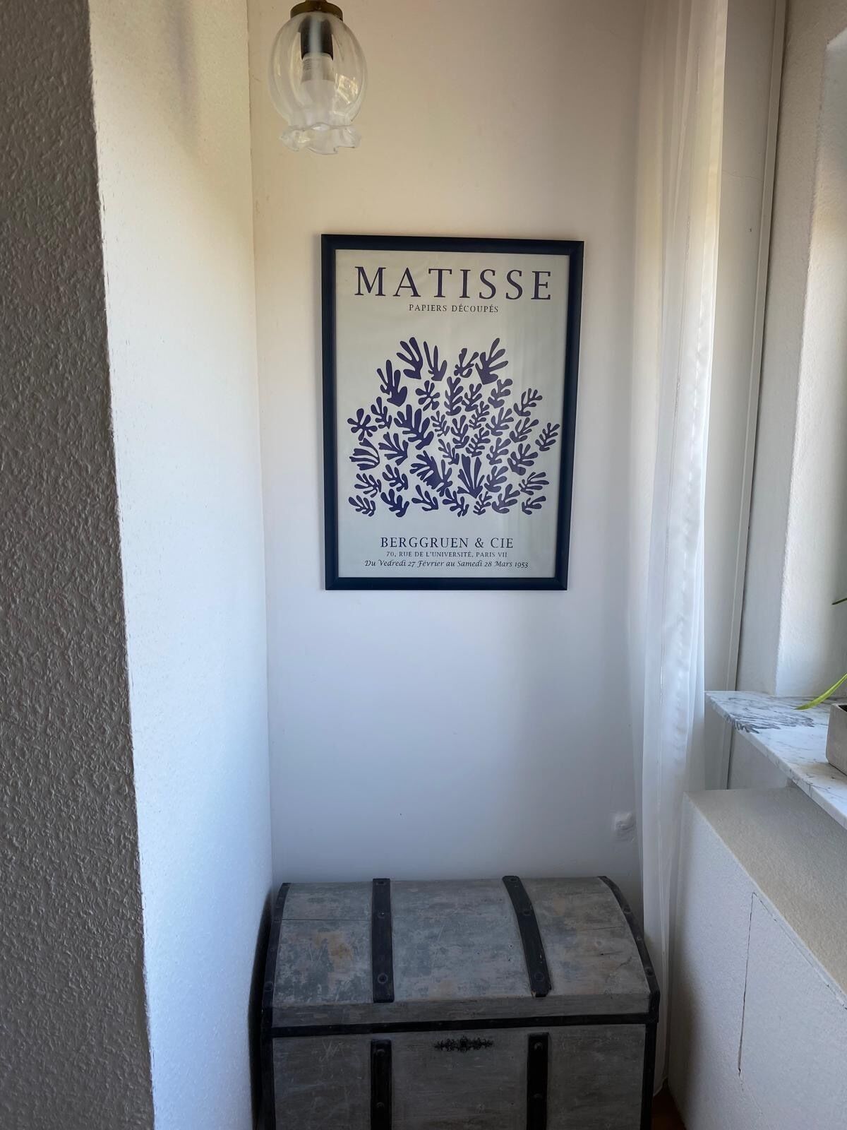 Una obra de arte enmarcada de una planta en la pared, un pequeño cofre de madera y una lámpara.