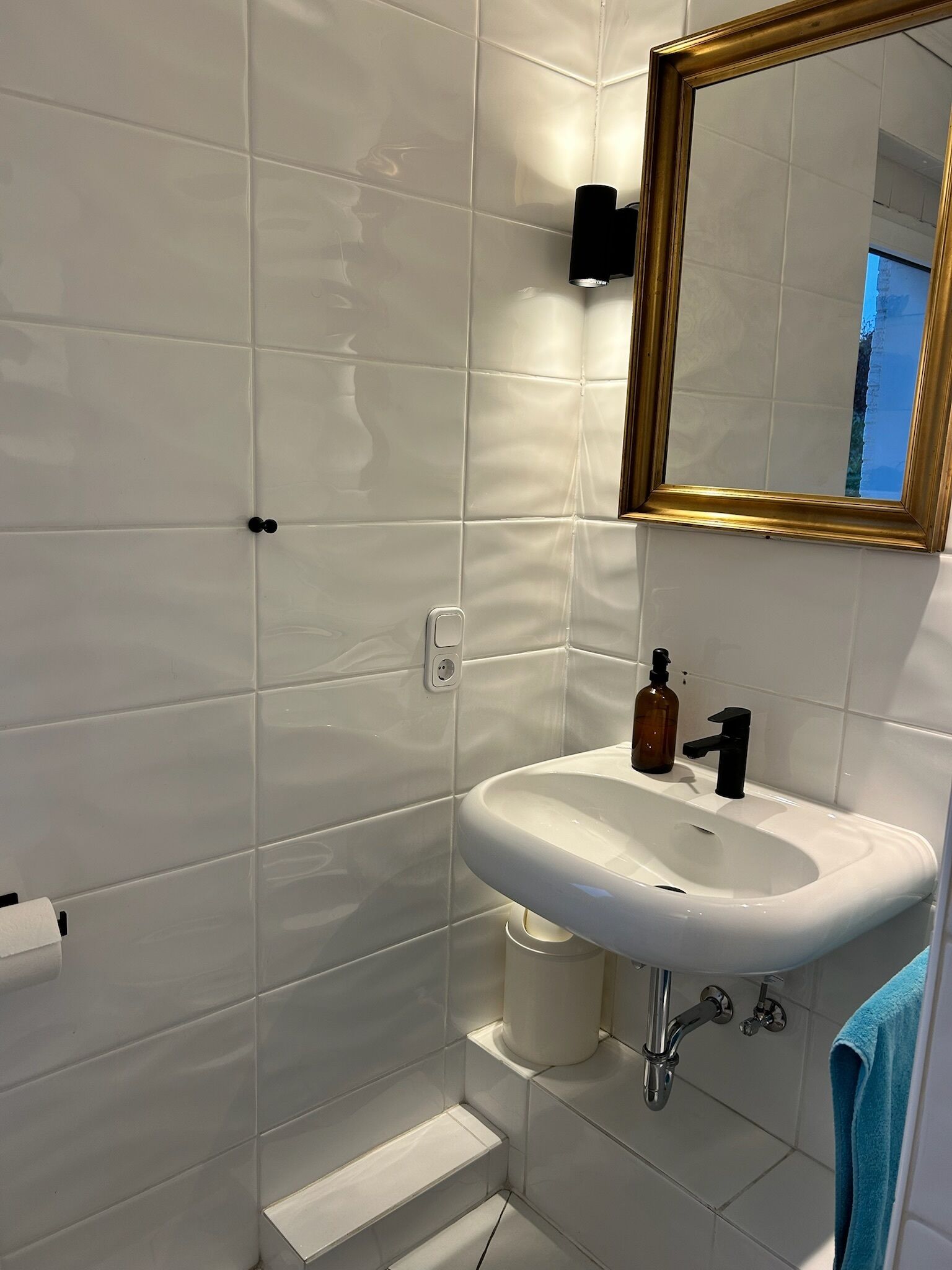Ein Badezimmer mit weißen Fliesen, einem weißen Waschbecken, einem schwarzen Wasserhahn, einem goldfarbenen Spiegelrahmen und einem blauen Handtuch.