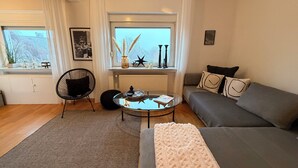 Ein gemütliches Wohnzimmer mit einer Couch, einem runden Couchtisch, einem Sessel und einem Fenster mit Vorhängen.