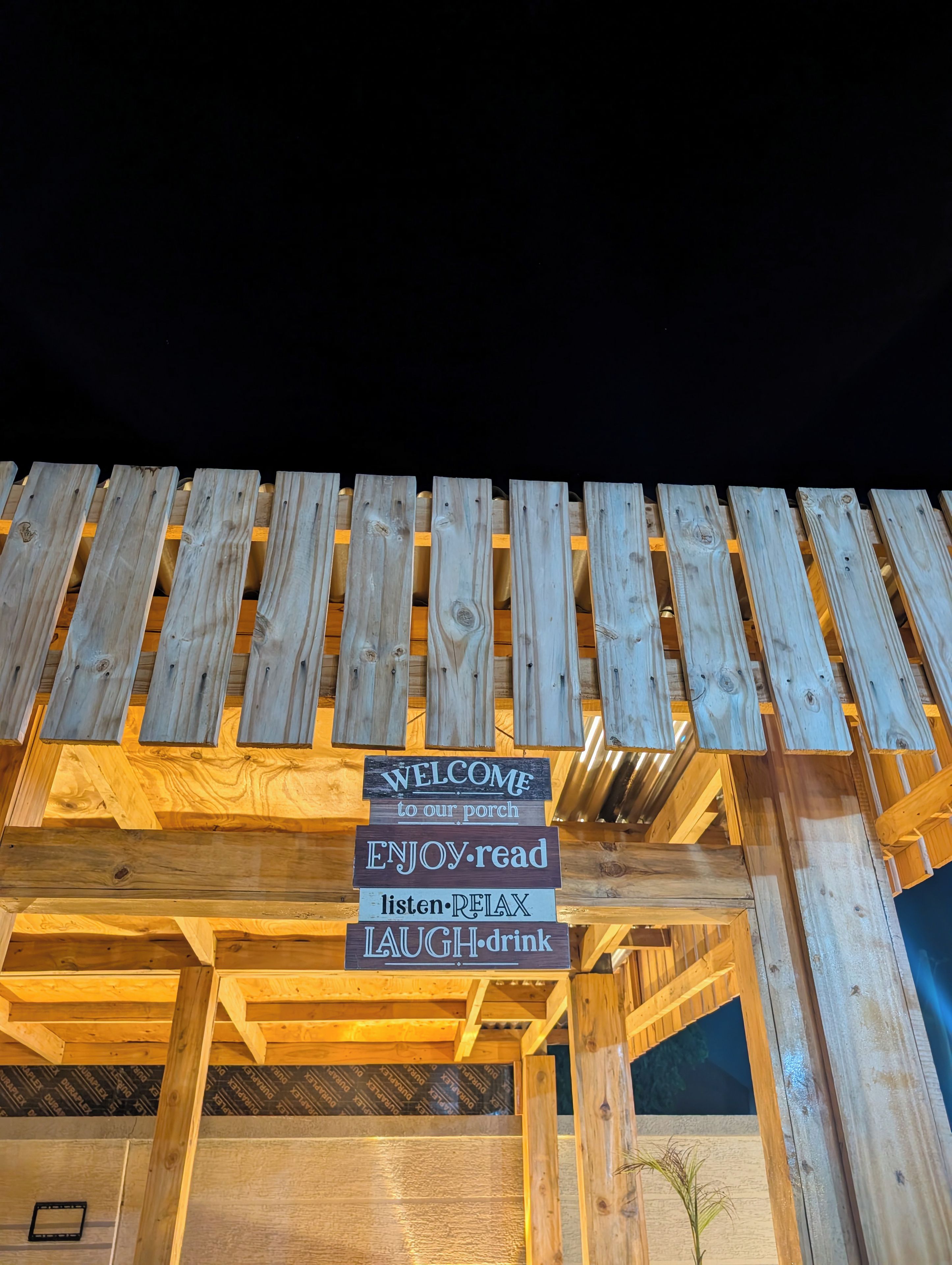 Una estructura de pérgola de madera con un letrero de bienvenida y una lista de actividades como leer, escuchar, relajarse, reír y beber.