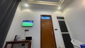 A room with a wooden door, a flat-screen TV mounted on the wall, a small table with a laptop and water bottles, and a ceiling light fixture. - La Million Palace (Dodoma)