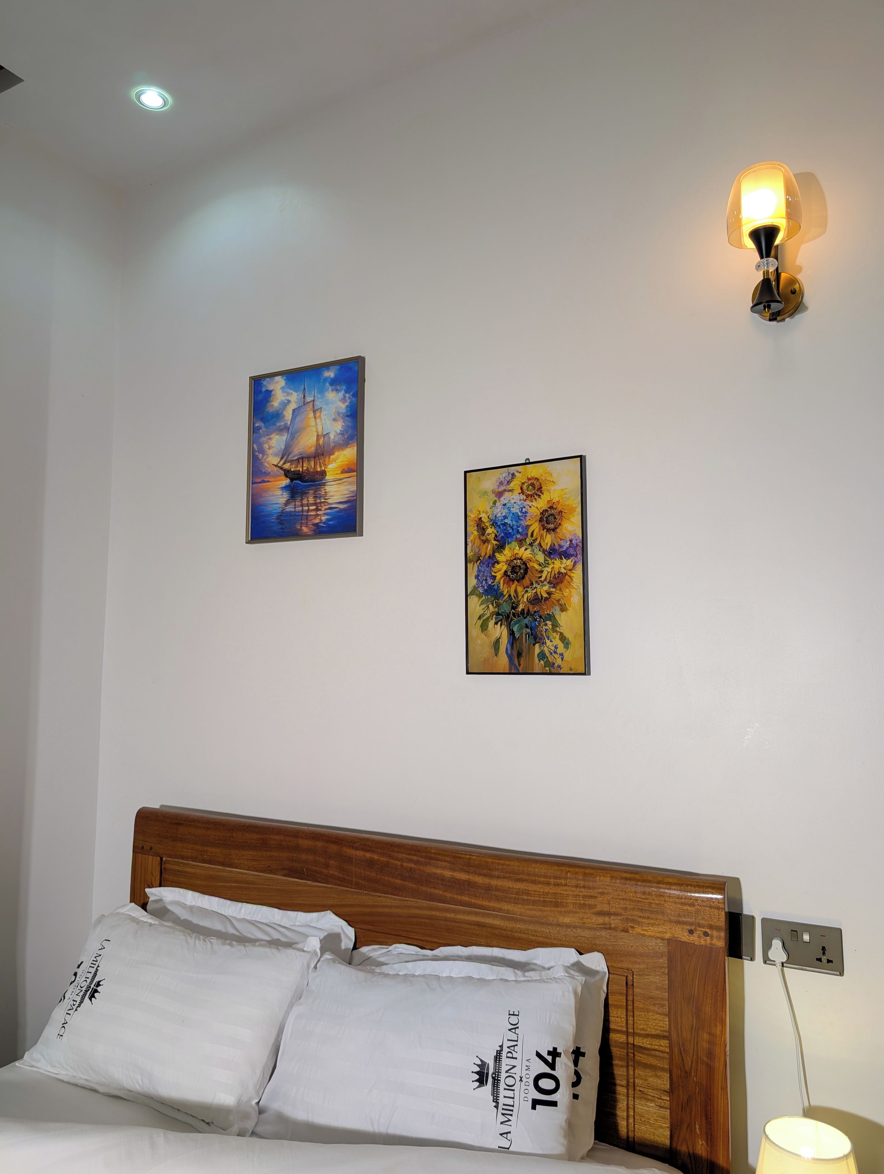 A hotel room with a wooden headboard, two white pillows, a painting of a sailboat, a painting of sunflowers, and a wall-mounted lamp.