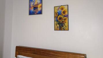 A hotel room with a wooden headboard, two white pillows, a painting of a sailboat, a painting of sunflowers, and a wall-mounted lamp.