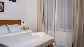 Una cama bien hecha con sábanas blancas y un cabecero decorativo, una mesita de noche con lámpara, y una ventana con cortinas traslúcidas.