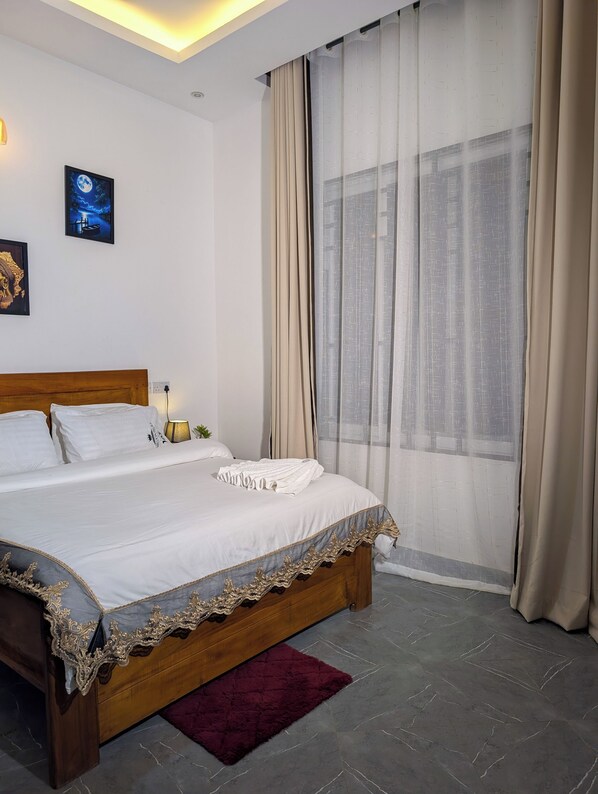 Una cama bien hecha con sábanas blancas y un cabecero decorativo, una mesita de noche con lámpara, y una ventana con cortinas traslúcidas.