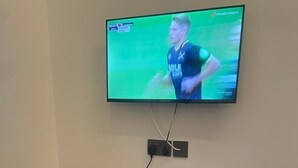A television mounted on a wall displaying a sports event, a wooden table with a laptop, a coffee cup, and a roll of toilet paper. - La Million Palace (Dodoma)
