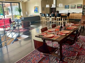 A dining area with a long table set for a meal, surrounded by chairs, with a kitchen in the background. - Provencal Villa With Garden & Pool (Uzès)