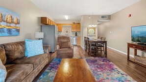 A living room with a brown sofa, a coffee table, and a colorful rug. There is a dining area with a table and chairs, and a kitchen in the background.