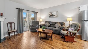 Ein Wohnzimmer mit Holzboden, einem grauen Sofa, zwei Sesseln, einem Couchtisch und einem Kamin.