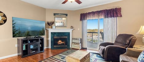 Una sala de estar con chimenea, ventilador de techo, sillón de cuero, un cofre de madera y televisión.