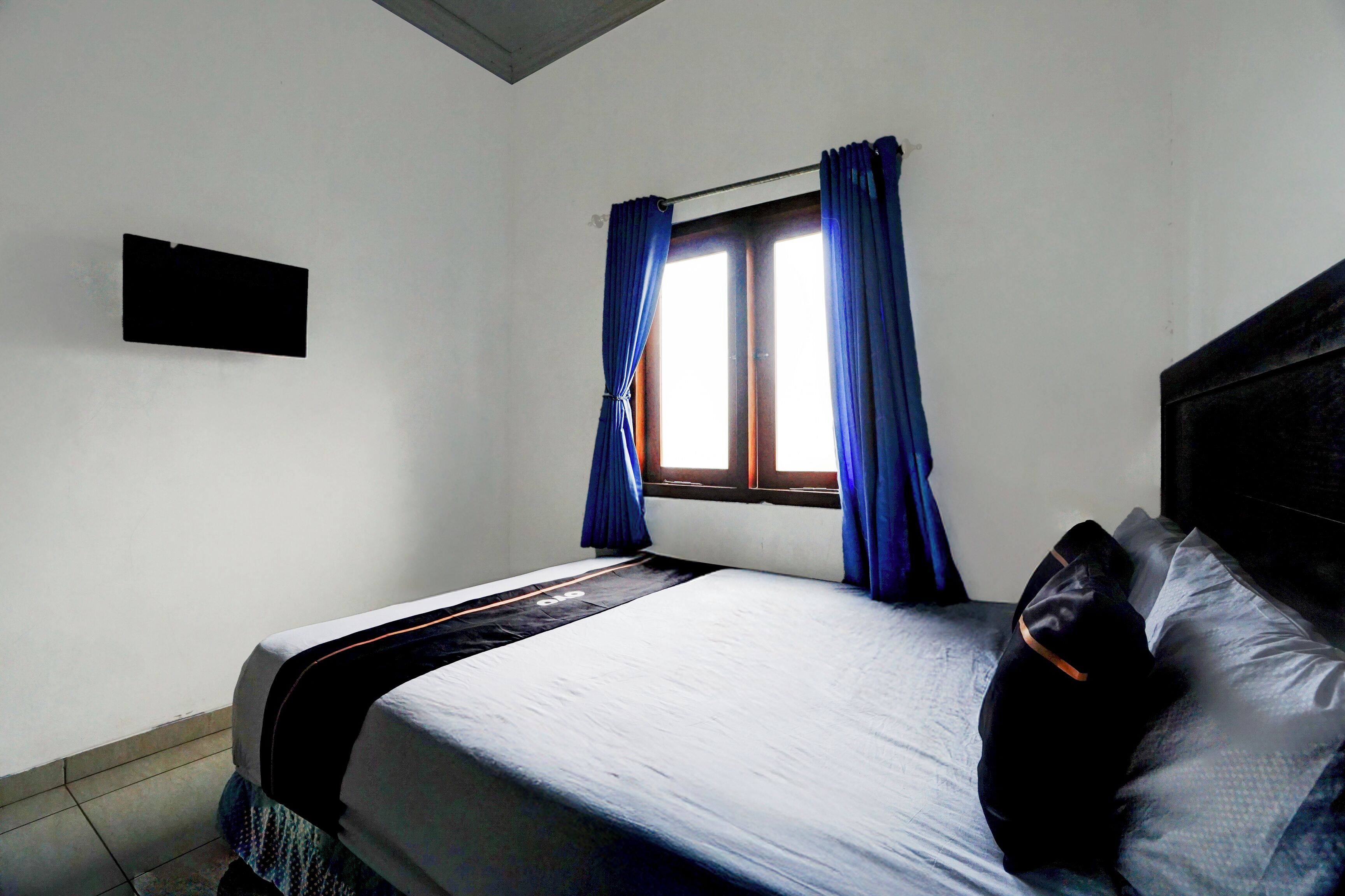 A single bed with a headboard, a window with blue curtains, and a wall-mounted TV.