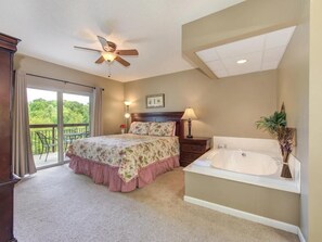 A bedroom with a large bed, a sitting area with a chair, a ceiling fan, a large window, and a bathtub.
