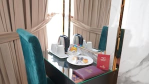 Hotelkamer met een tafel met glazen blad, een spiegel, een theeset, een bord met snacks en een rood boekje.