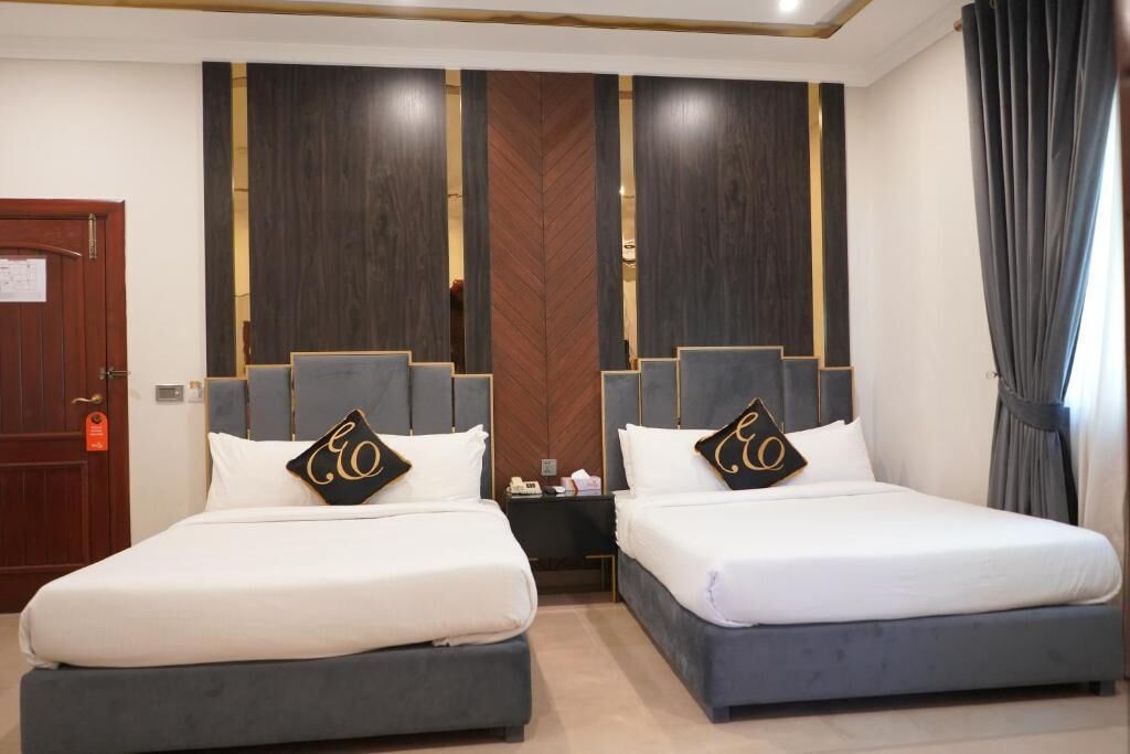 A hotel room with two beds, each with a grey headboard and white bedding. There is a wooden door on the left and a curtain on the right.