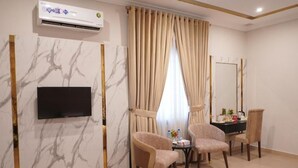 Hotelkamer met een flatscreen televisie aan de muur, een wand met marmerpatroon, een groot raam met beige gordijnen, twee fauteuils, een kleine ronde tafel en een bed met een witte dekbedovertrek.