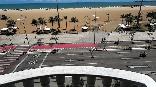 Blick auf einen Strand vom Balkon aus, mit Palmen, einer Straße mit roter Fahrradspur und blauem Himmel.