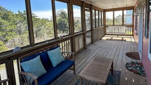 Un porche cubierto con un banco de madera y una mesa, ventanas amplias y vistas a árboles.