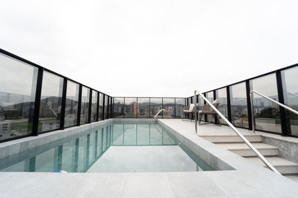 Une piscine sur le toit, entourée de garde-corps en verre, un espace détente avec des fauteuils et une vue sur le paysage urbain.
