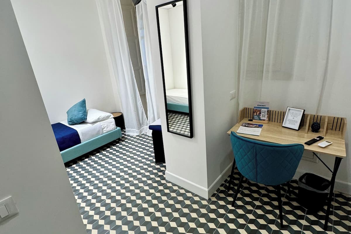 A room with a checkered floor, a bed with blue bedding, a desk with a chair, and a mirror on the wall.