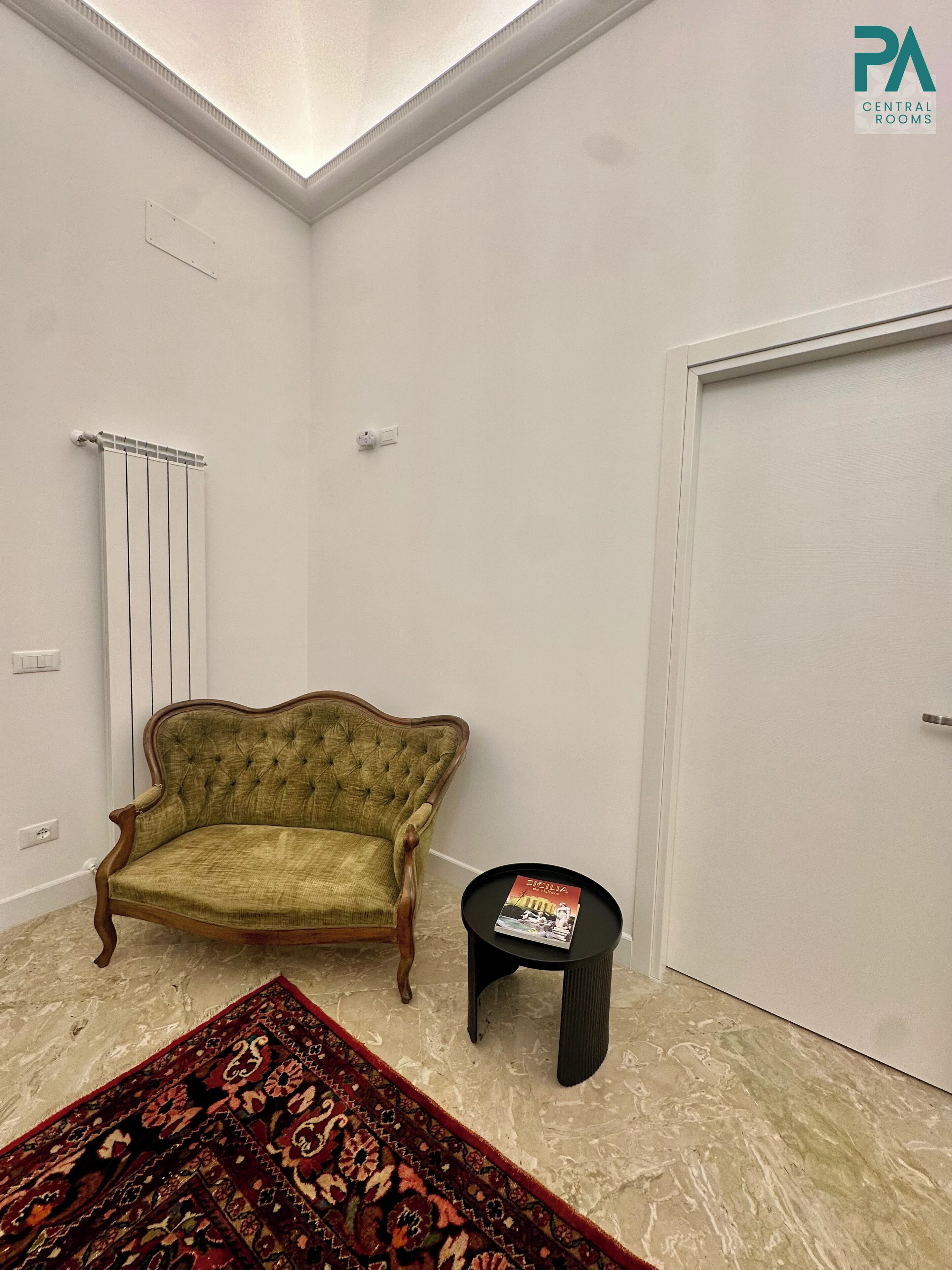 Una habitación con un sillón tapizado en verde, una pequeña mesita auxiliar con un libro, una alfombra estampada, un radiador y una puerta cerrada.